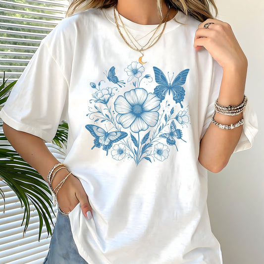 White t-shirt with blue floral and butterfly design worn by a person, with a neutral background.