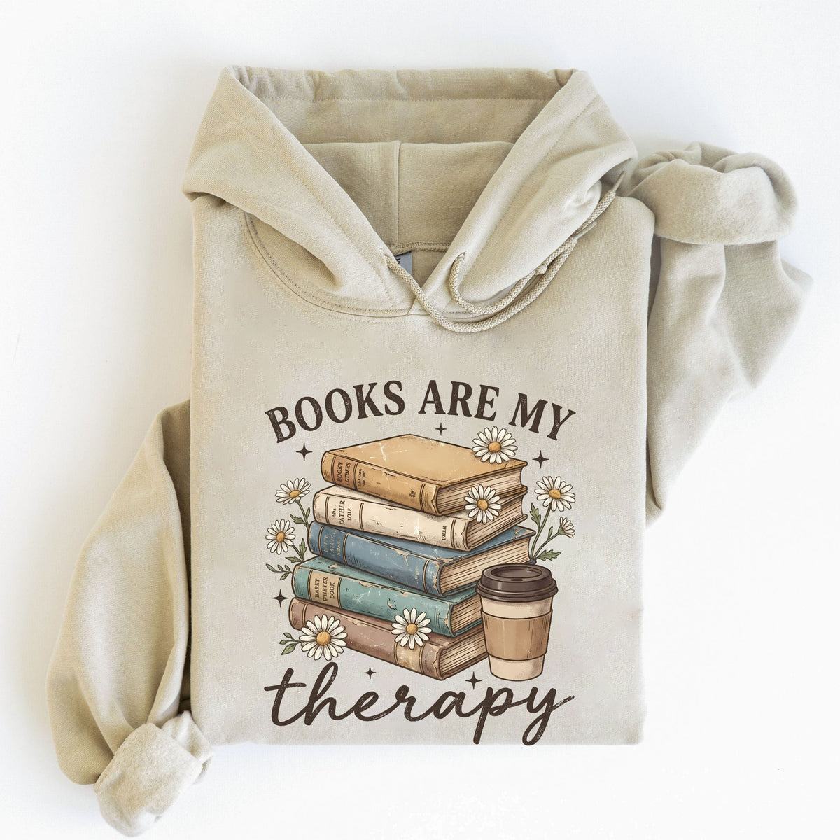 A beige hoodie laid flat on a white background, featuring a graphic print of a stack of books, small daisies, and a coffee cup with the text 'BOOKS ARE MY therapy'.