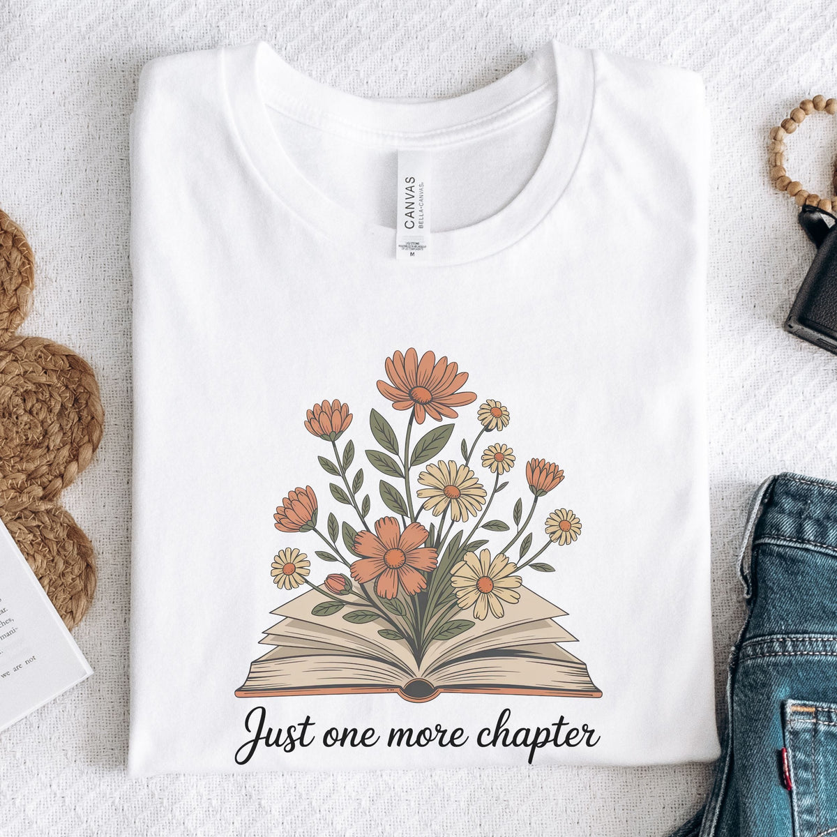 A folded white graphic T-shirt on a light textured surface, featuring an illustration of an open book with orange, yellow, and white flowers, and the text 'Just one more chapter' printed below. A Canvas neck label is visible, and styled props including woven mats, jeans, and a watch are partially visible around the shirt.