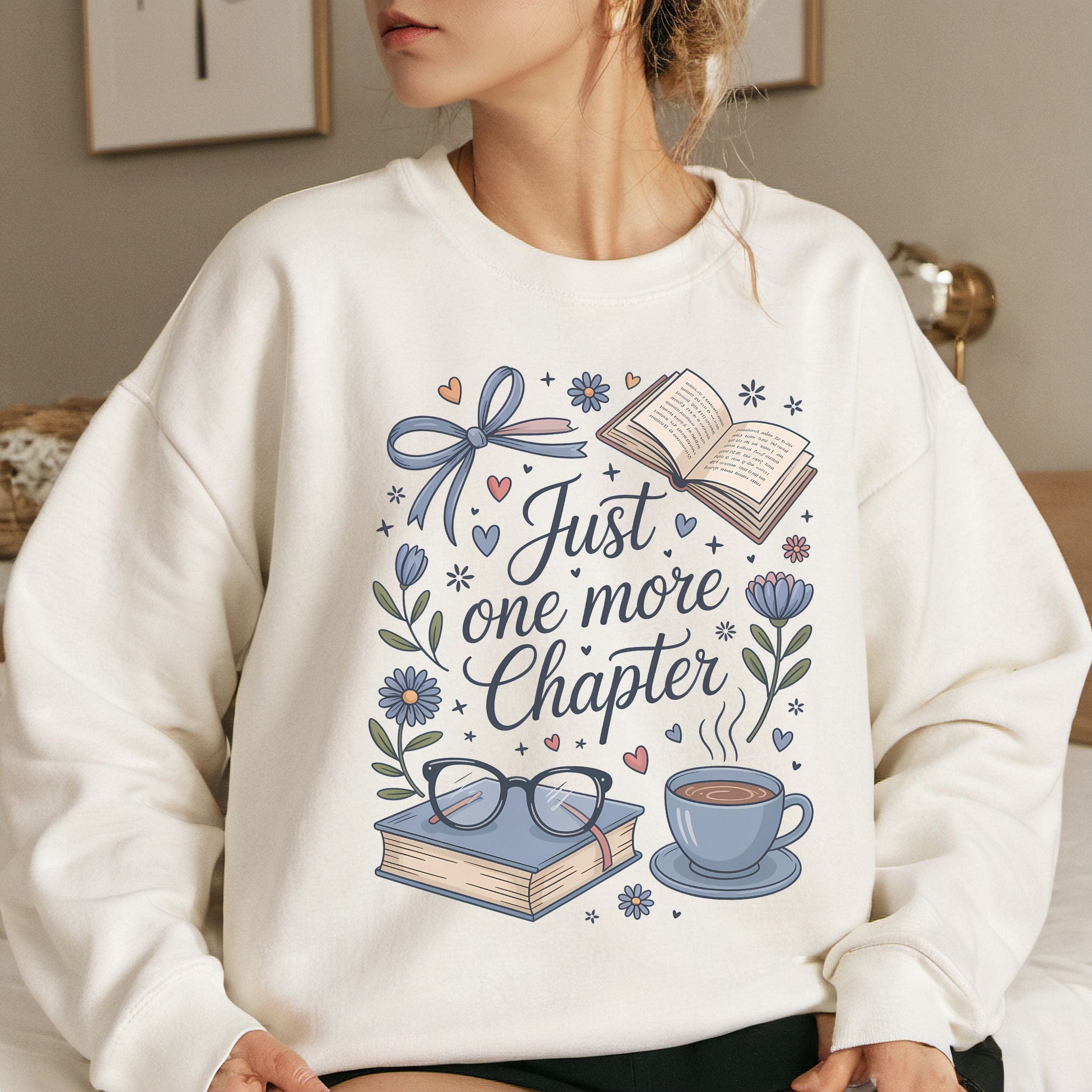Person wearing a white sweatshirt with 'Just one more chapter' design featuring books, glasses, and a cup.