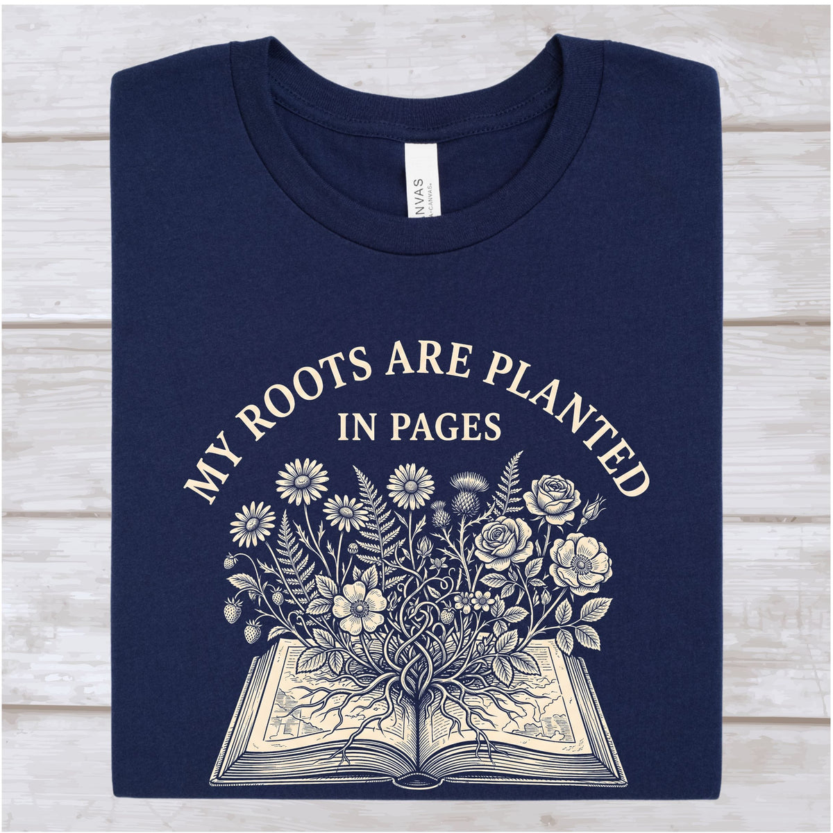 A navy blue crewneck t-shirt with a cream-colored graphic print that reads 'MY ROOTS ARE PLANTED IN PAGES' above an illustration of flowers growing from an open book, laid flat on a light wood background.