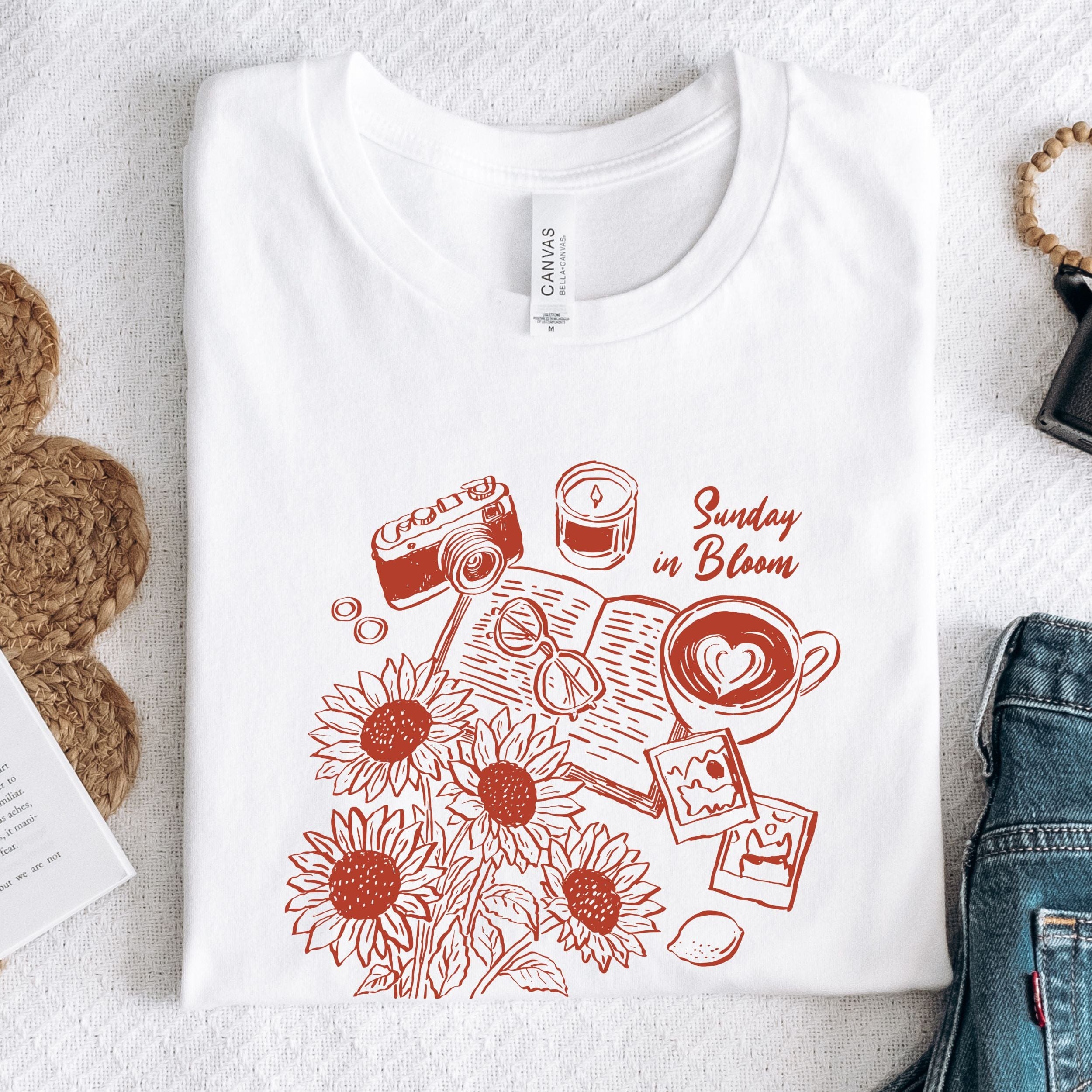White t-shirt with red graphic design featuring sunflowers, a camera, and text on a light gray surface.