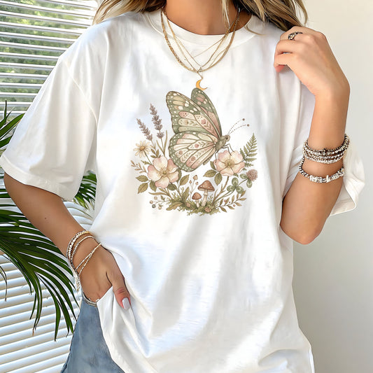 White t-shirt with a butterfly and floral design worn by a person, with a plant in the background.