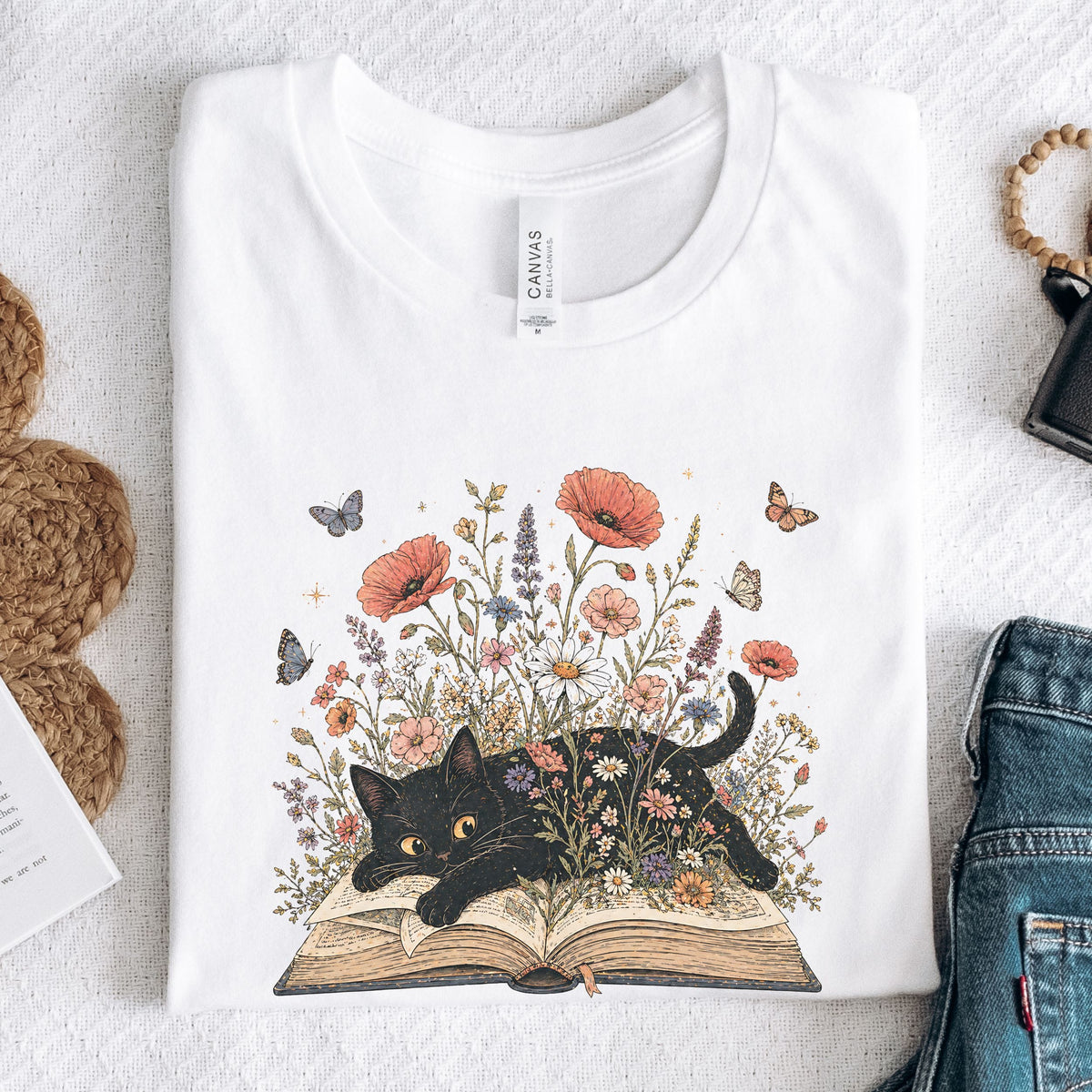 A folded white graphic T-shirt on a textured white surface, featuring a black cat lying on an open book surrounded by colorful wildflowers and butterflies. Styled flat-lay props include denim jeans, a woven bag, and a small card.