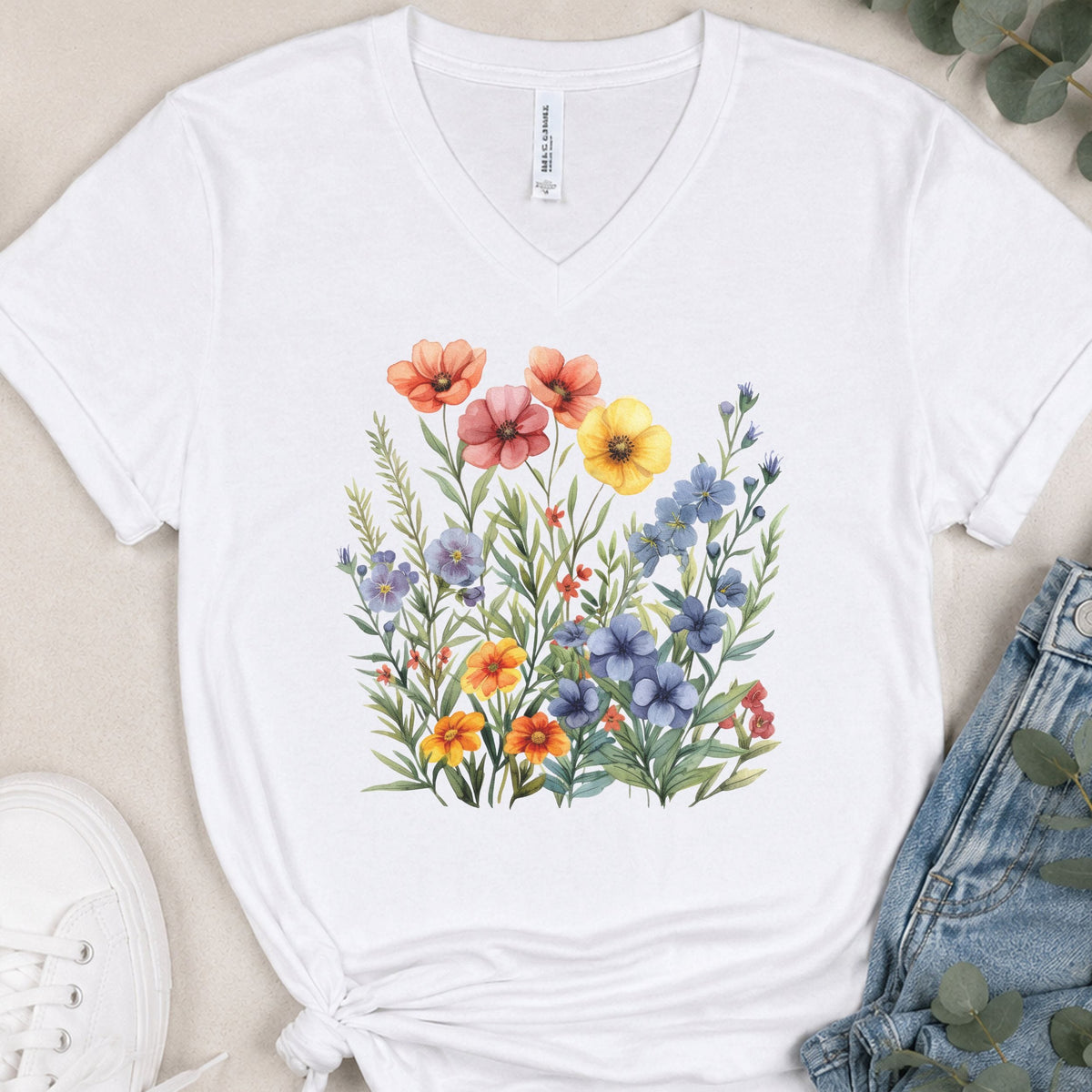 A white V-neck t-shirt with a colorful watercolor wildflower print on the front, styled in a flat lay with blue jeans, a white sneaker, and eucalyptus leaves on a beige background.
