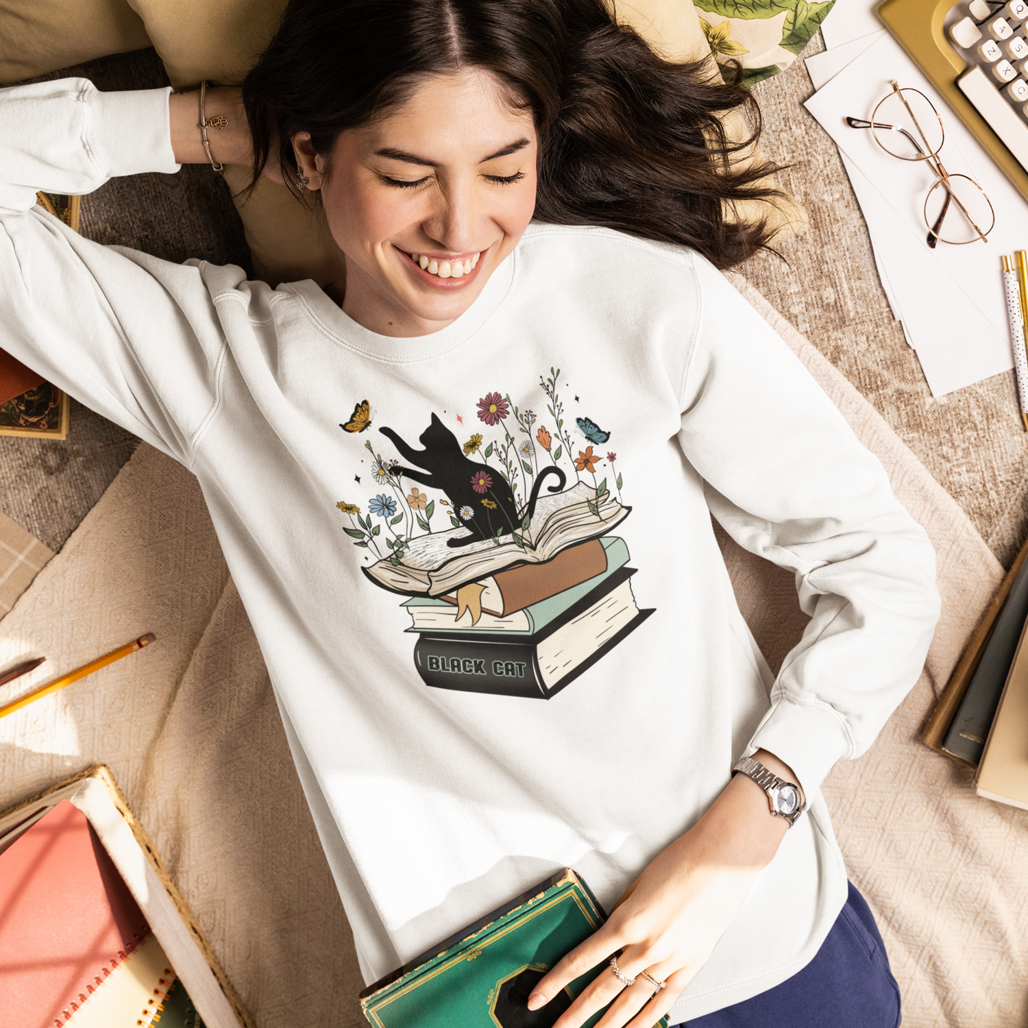 Cozy reading sweatshirt featuring a black cat, stacked books, and botanical floral artwork, styled in a warm and inviting book lover setting.