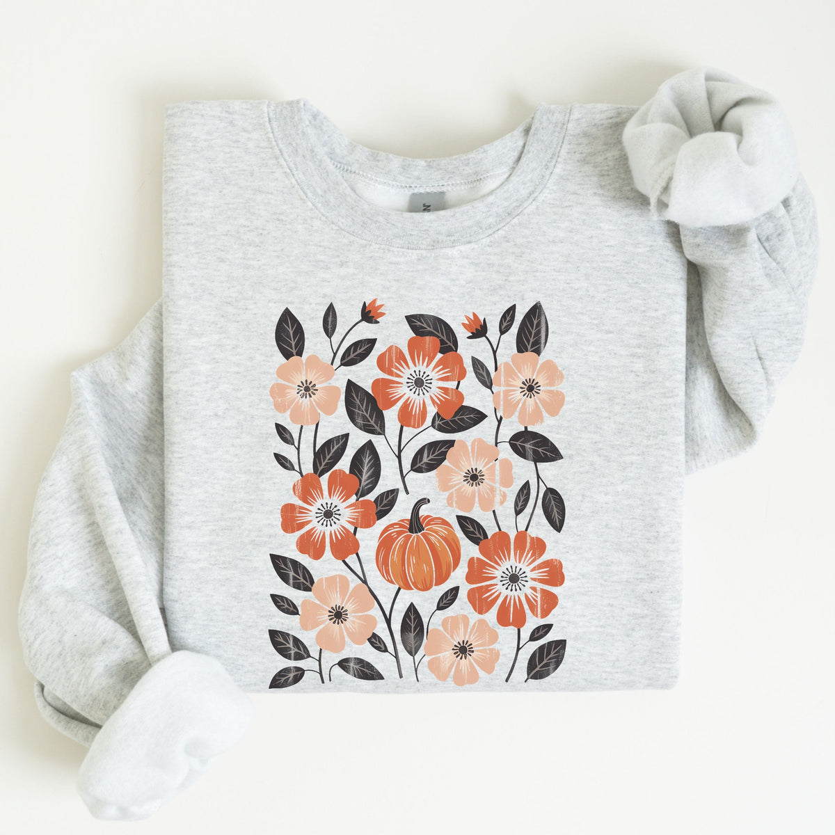 A folded light heather gray sweatshirt featuring a central autumn floral and pumpkin graphic in orange, peach, and dark gray, styled with a soft white scrunchie on a warm off-white background.