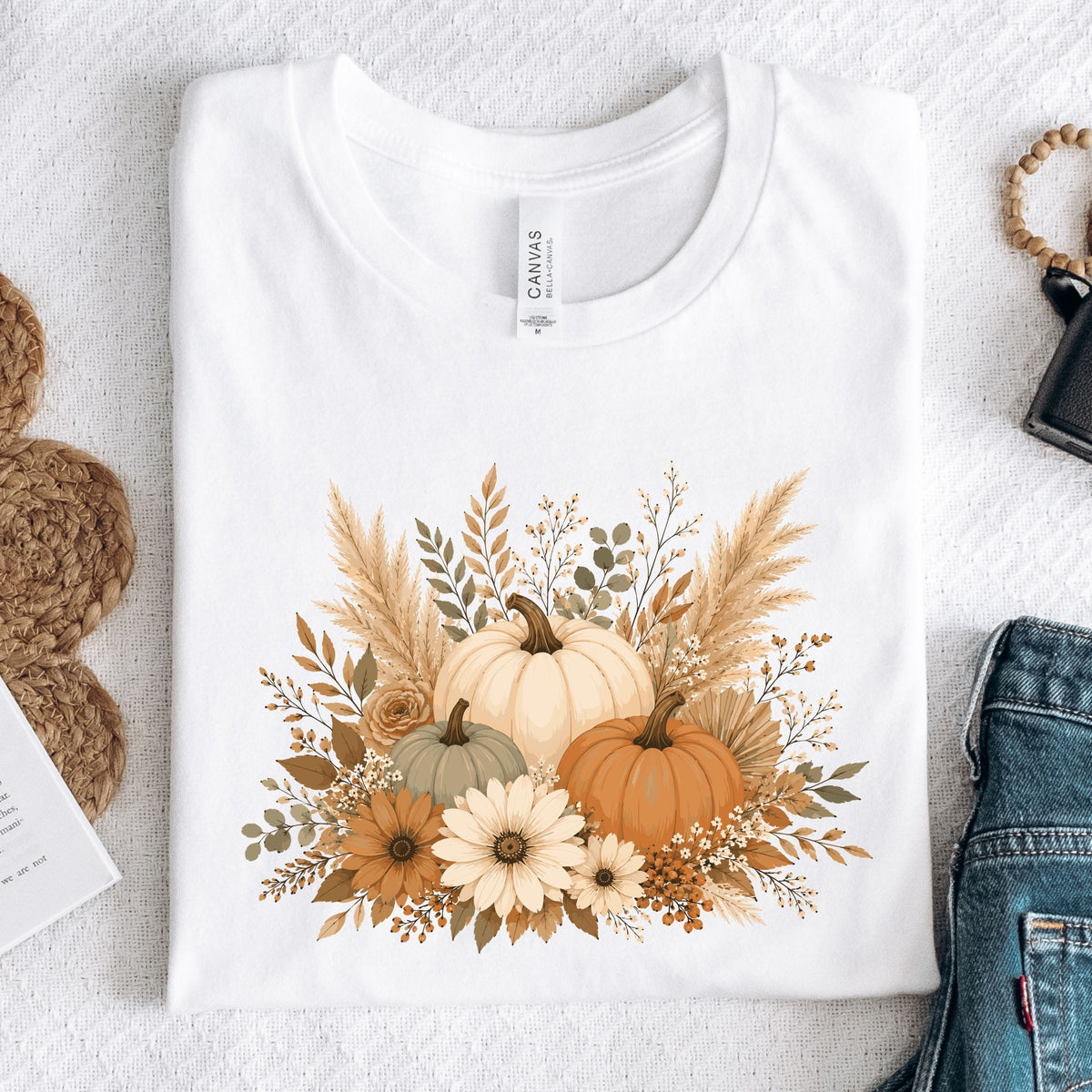 A folded white long sleeve T-shirt with a fall floral pumpkin graphic, styled in a flat lay on a textured white surface with jeans, a woven bag, and a small card nearby. The neck label reads Canvas.
