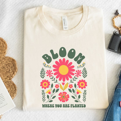 Beige t-shirt with floral design and 'Bloom Where You Are Planted' text on a textured surface.