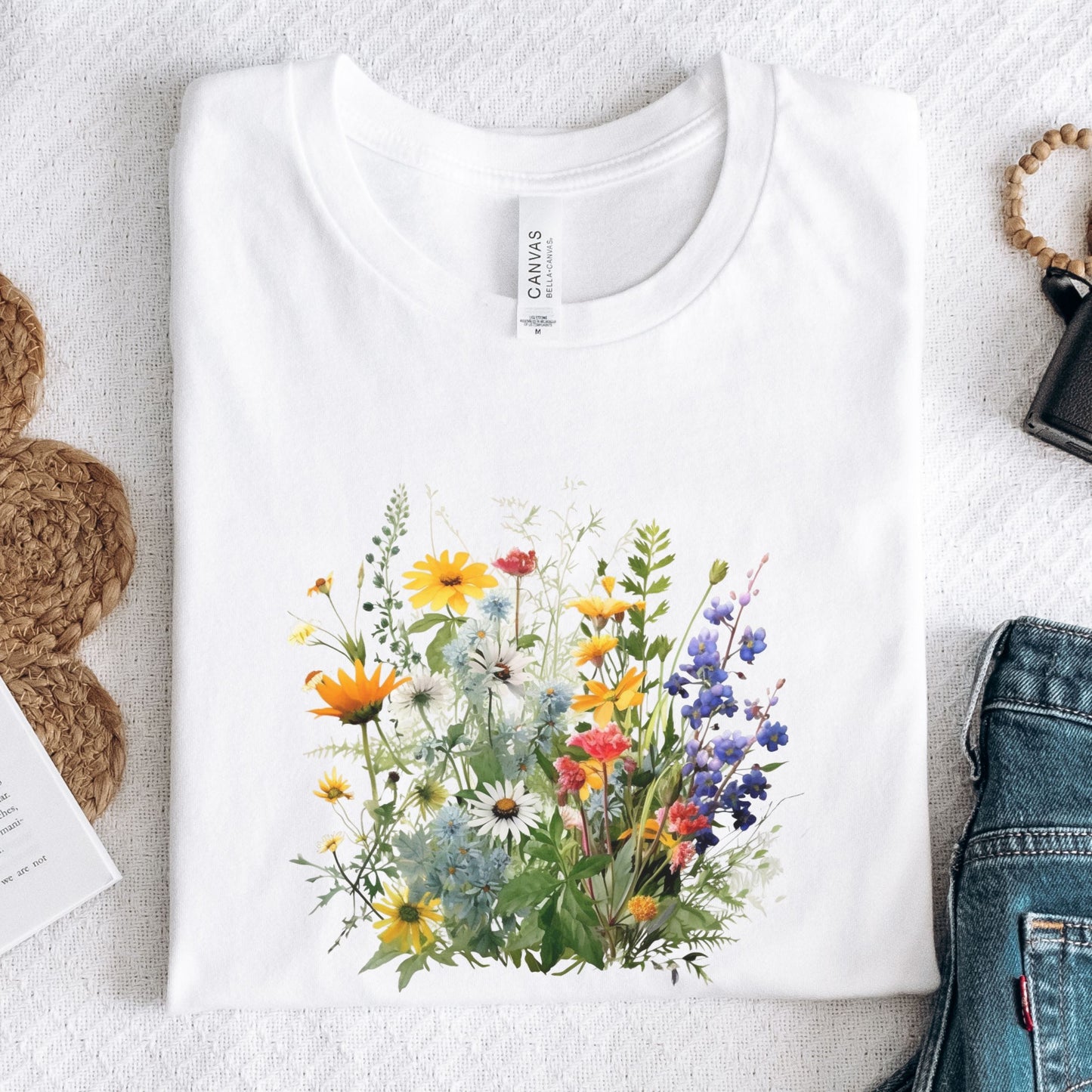 A folded white t-shirt with a colorful wildflower graphic print, displayed in a flat-lay arrangement on a textured white background with jeans, a woven bag, and a book partially visible.