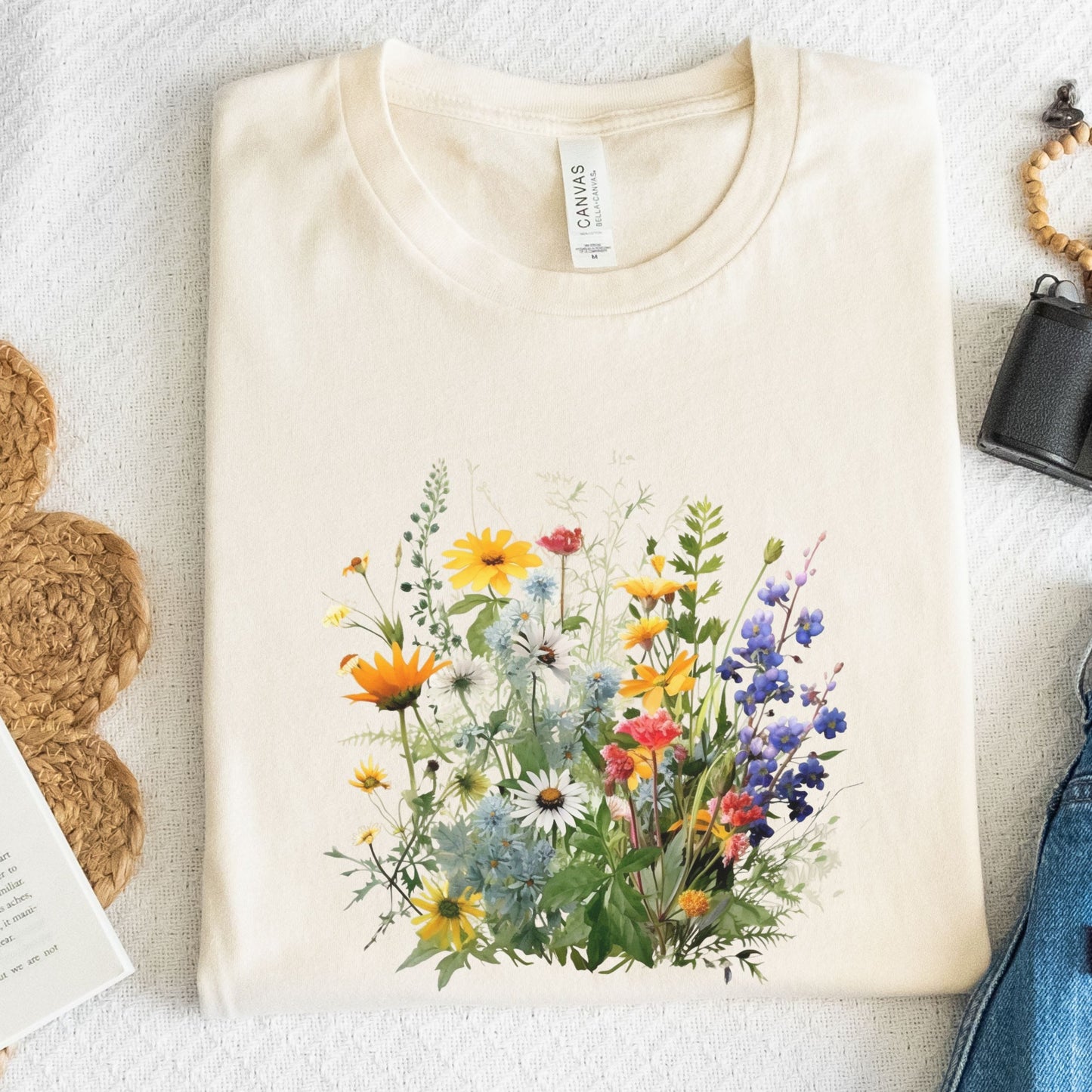 A folded beige t-shirt with a colorful wildflower graphic print, displayed in a flat-lay arrangement on a textured white background with jeans, a woven bag, and a book partially visible.