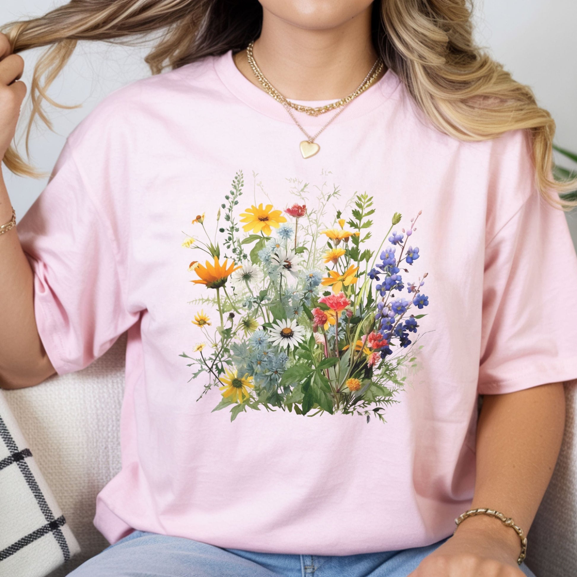 A woman wearing a light pink short-sleeve t-shirt with a colorful watercolor-style wildflower graphic on the front, paired with blue jeans and gold jewelry.