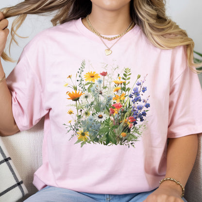 A woman wearing a light pink short-sleeve t-shirt with a colorful watercolor-style wildflower graphic on the front, paired with blue jeans and gold jewelry.