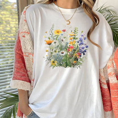 A woman wearing a white oversized T-shirt with a colorful wildflower graphic on the front, layered with a coral and cream knit cardigan. The background features a window with blinds and green plants.