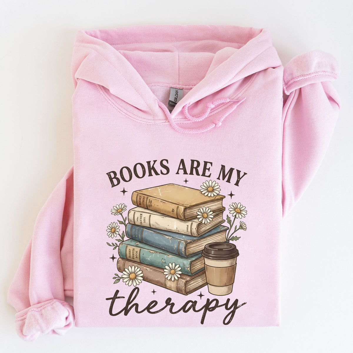 A light pink pullover hoodie laid flat on a white background, featuring a graphic print of a stack of books, daisies, and a coffee cup with the text 'BOOKS ARE MY therapy'.