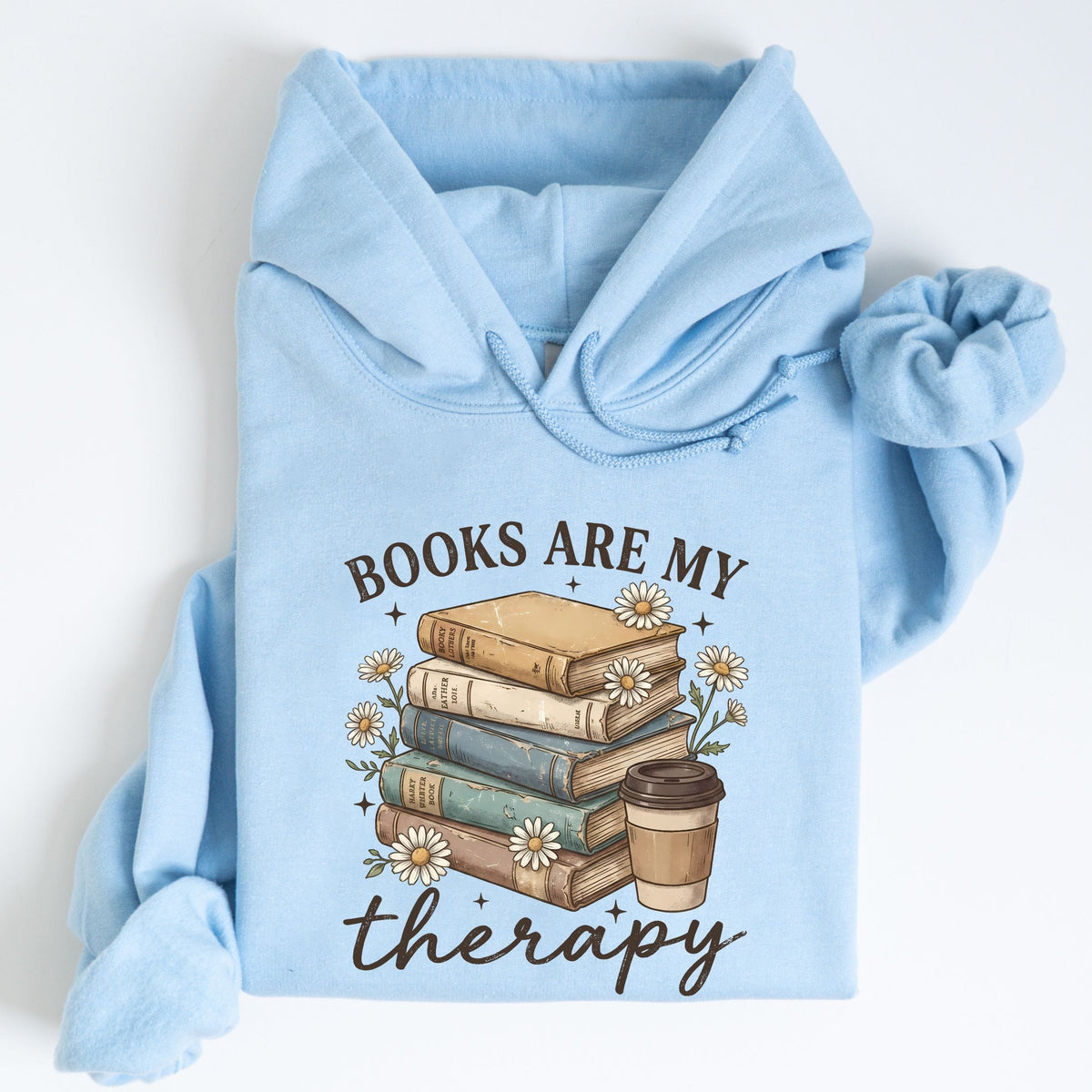 A light blue hoodie laid flat on a white background, featuring a graphic print of a stack of books, small white flowers, and a takeaway coffee cup with the text 'BOOKS ARE MY therapy'.