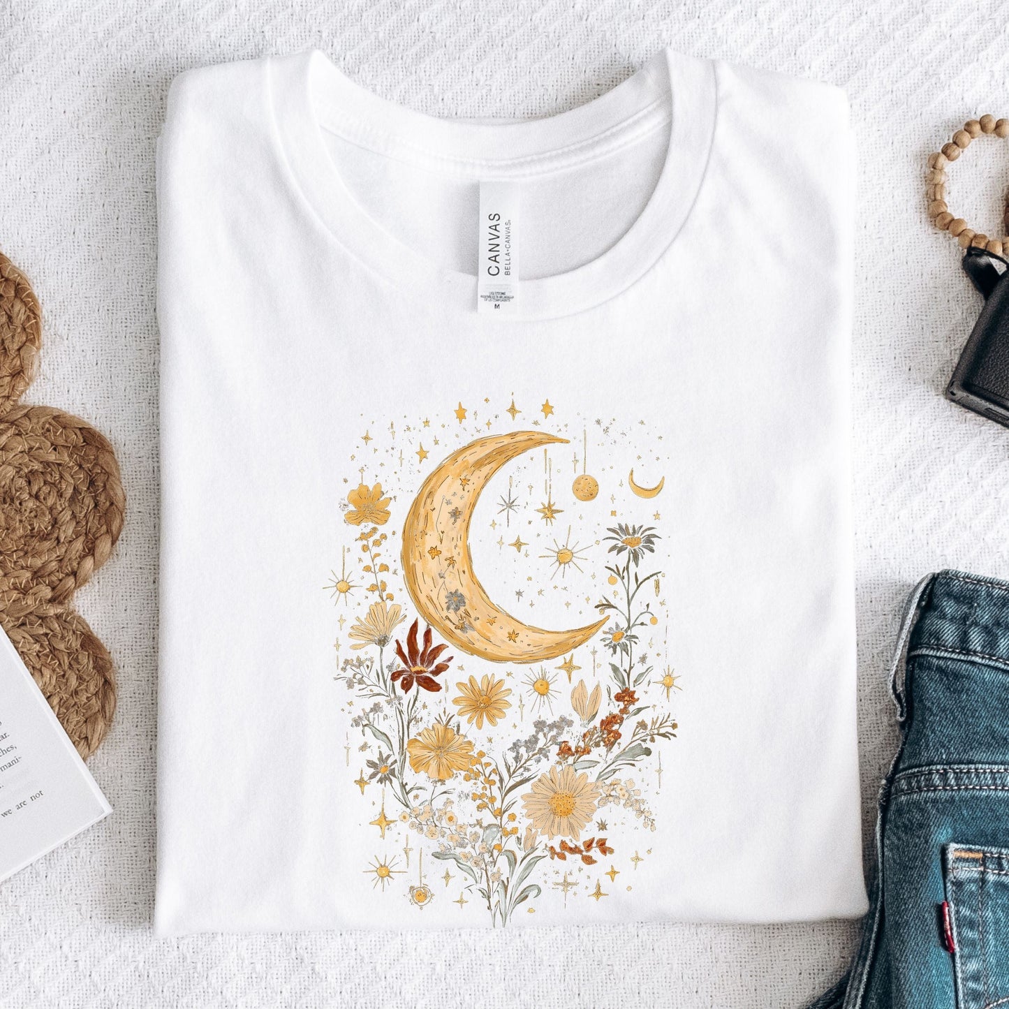 White t-shirt with a decorative moon and floral design on a textured surface.