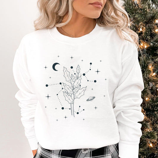 Person wearing a white sweatshirt with black leaf design and celestial elements, standing next to a decorated Christmas tree.