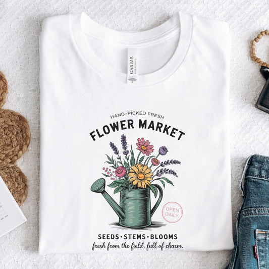 White t-shirt with 'Flower Market' graphic design featuring a watering can and flowers on a textured surface.