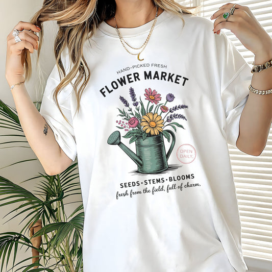 White t-shirt with 'Flower Market' graphic design worn by a person, with plants in the background.