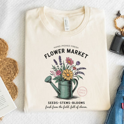 Beige t-shirt with 'Flower Market' graphic design featuring a watering can and flowers on a textured surface.