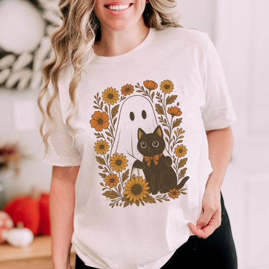 Person wearing a white t-shirt with a Halloween-themed design featuring a ghost, black cat, and flowers.