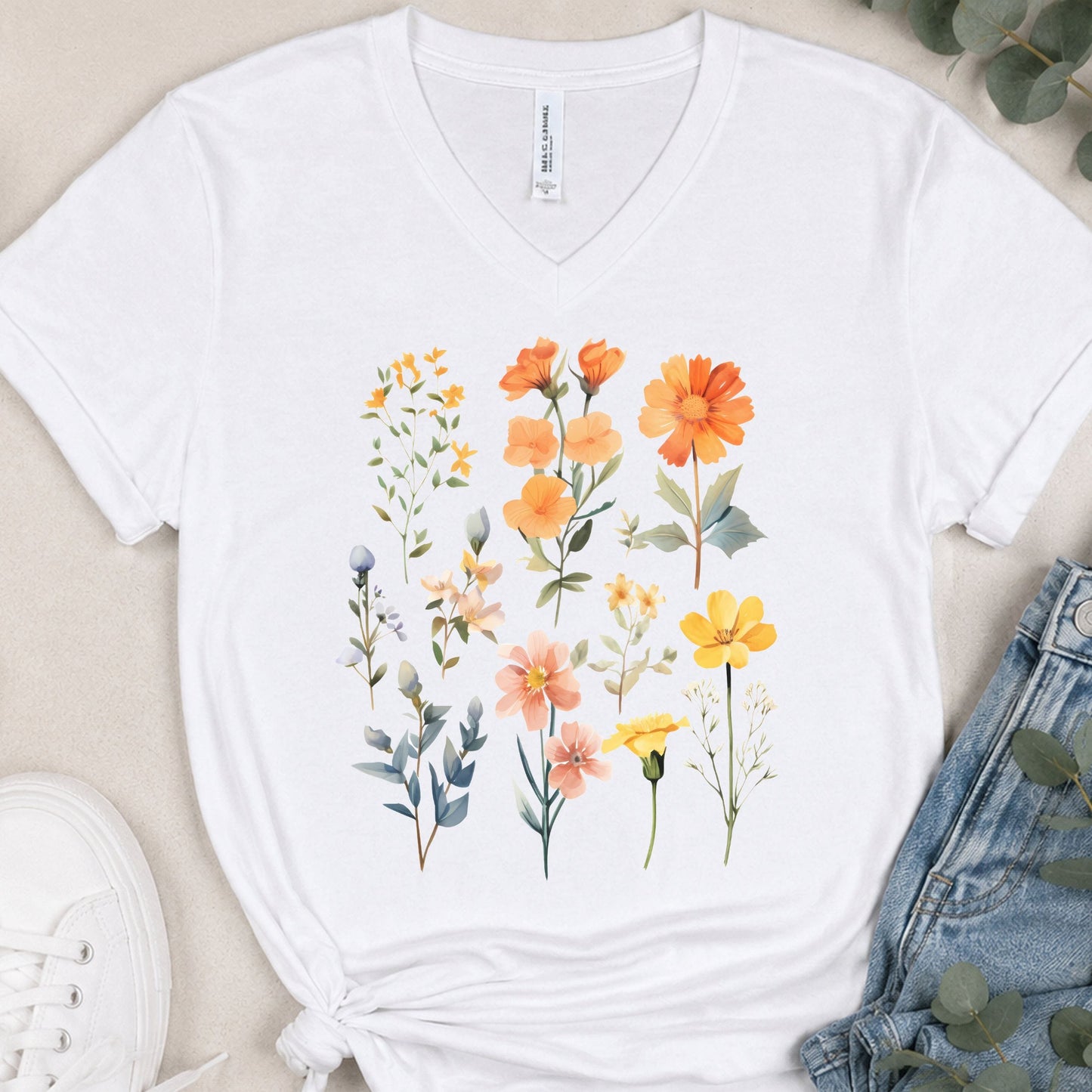 A white V-neck t-shirt with a colorful watercolor floral print, styled in a flat lay with blue jeans, a white sneaker, and eucalyptus leaves on a beige background.