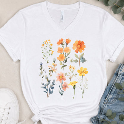 A white V-neck t-shirt with a colorful watercolor floral print, styled in a flat lay with blue jeans, a white sneaker, and eucalyptus leaves on a beige background.