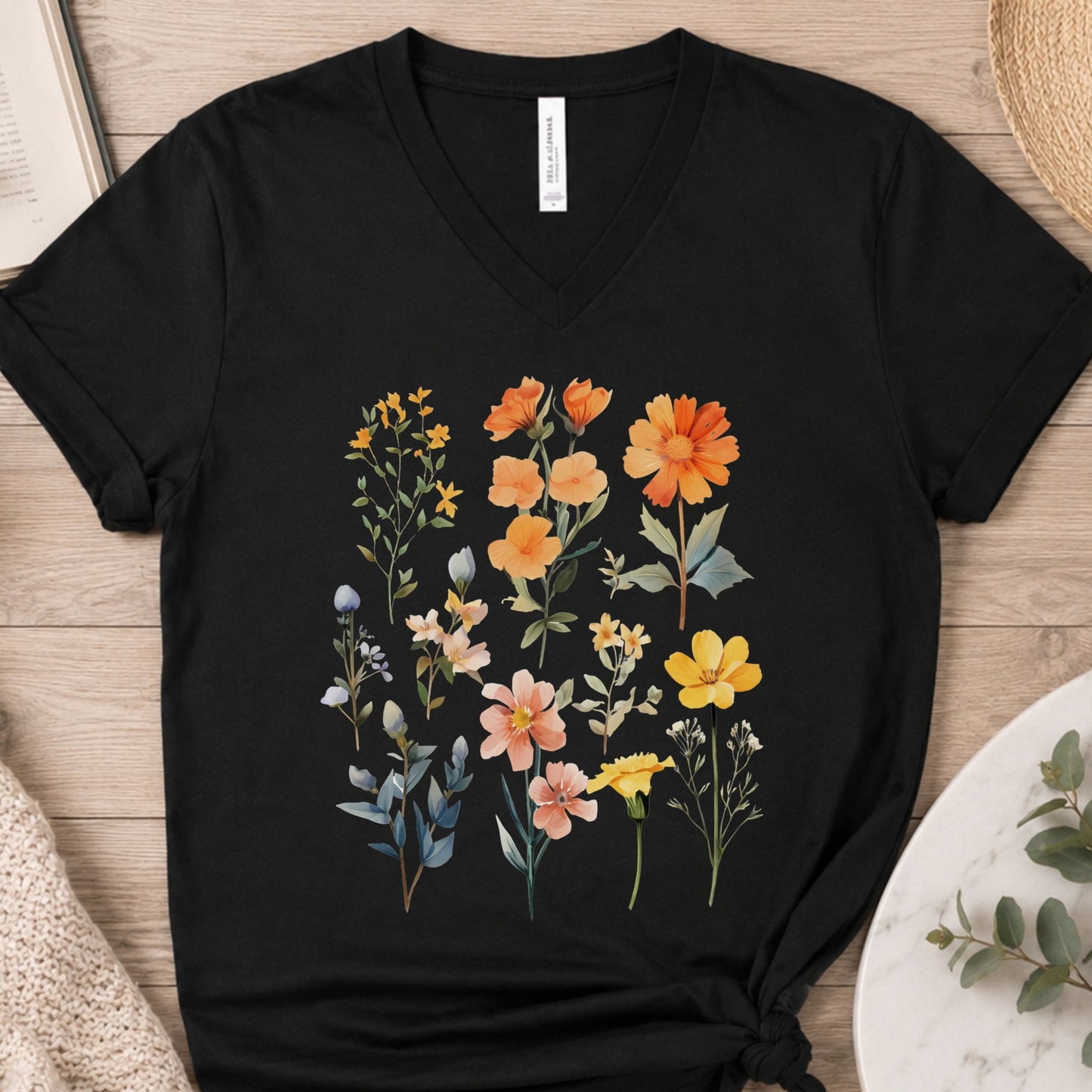 A black V-neck t-shirt with a colorful watercolor-style floral print, laid flat on a light wood background with styled props.