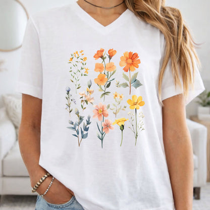 A woman wearing a white V-neck t-shirt with a colorful watercolor floral print of orange, yellow, and peach flowers with green leaves, set against a bright, softly blurred living room background.