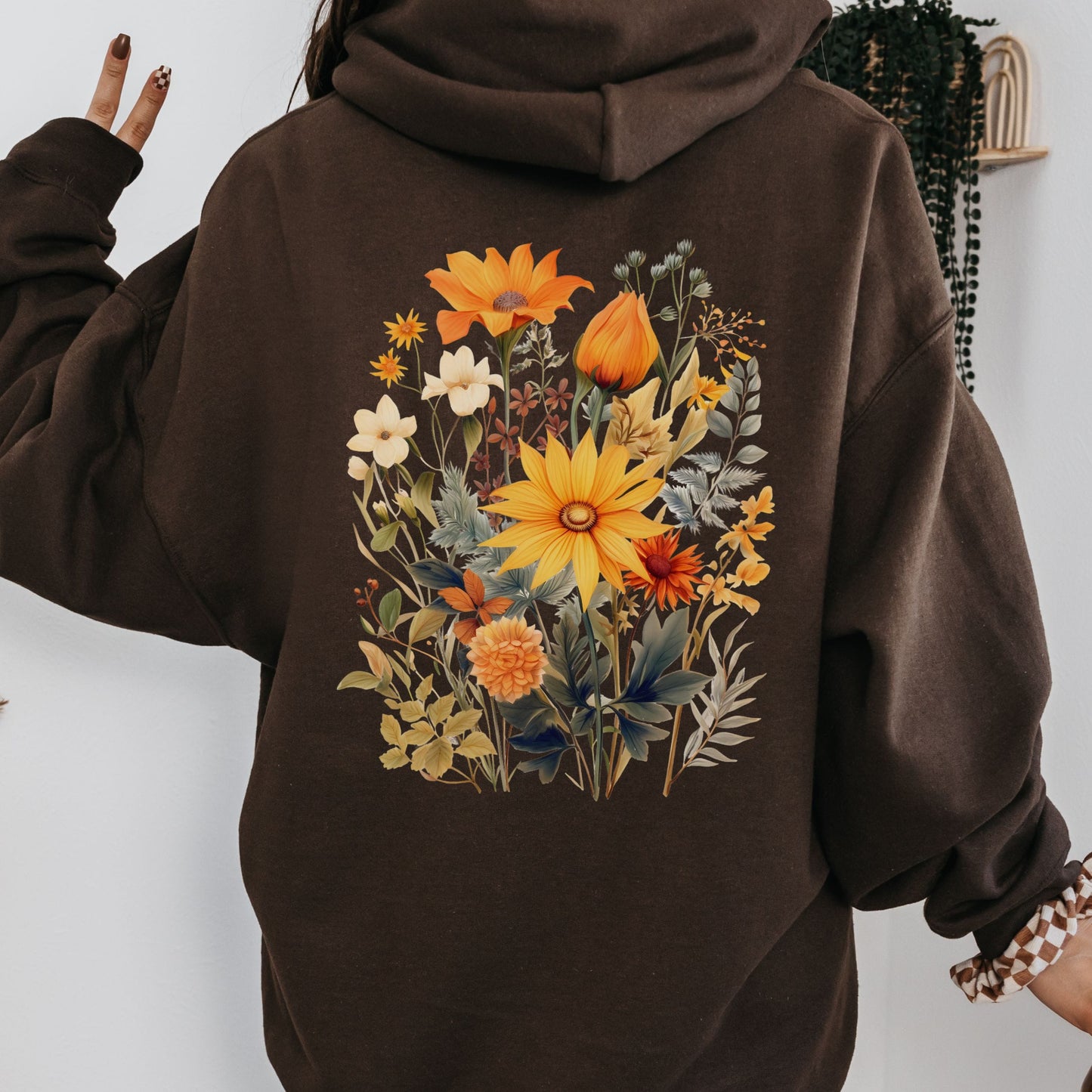 A person wearing an oversized dark brown hoodie with a large colorful floral print on the back, posed against a light wall with a hanging plant in the background.