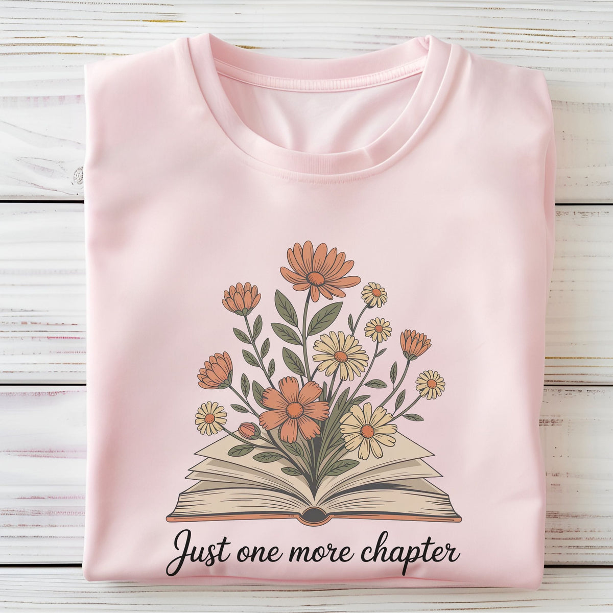 A folded light pink crewneck t-shirt on a white wood plank background, featuring a graphic of an open book with orange and yellow flowers growing from it, and the text 'Just one more chapter' printed below.