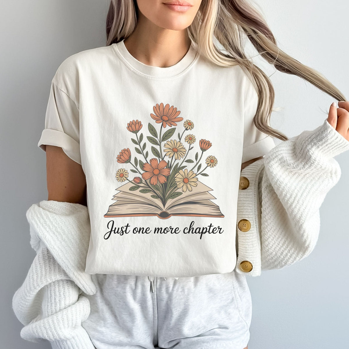 A woman wearing a cream-colored graphic t-shirt with a floral book design and the text 'Just one more chapter', styled with a chunky white knit cardigan and light gray sweatpants against a neutral gray background.