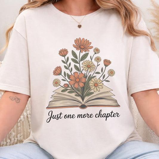 A woman wearing a cream-colored t-shirt with a graphic of an open book and flowers, with the text 'Just one more chapter' printed below. The photo is cropped from the chest down, showing the fit of the shirt.
