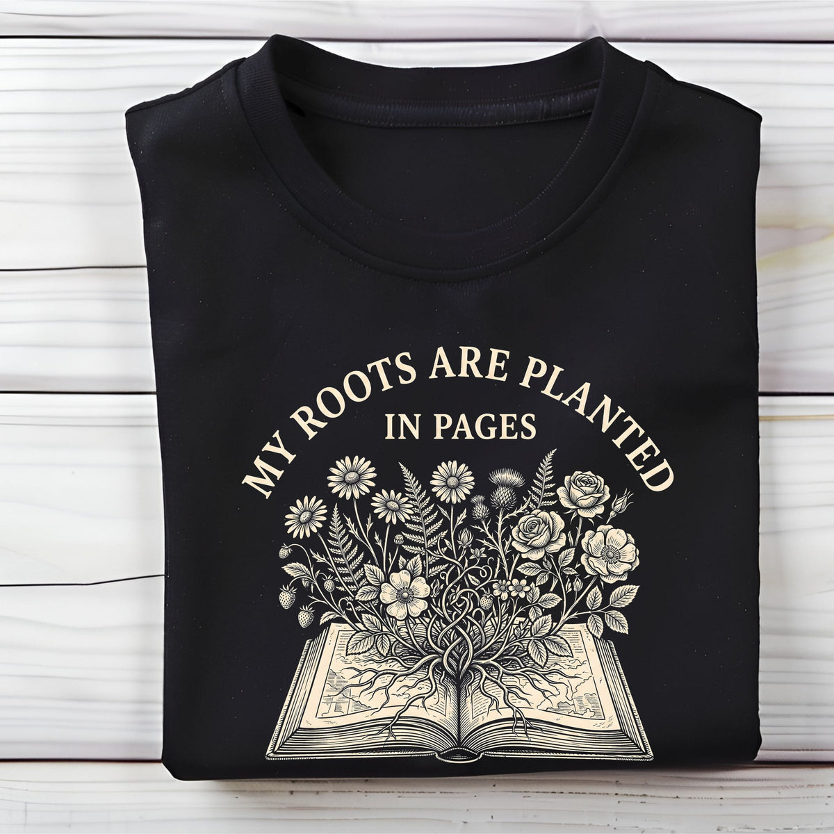 A folded black t-shirt with a cream-colored graphic print that reads 'MY ROOTS ARE PLANTED IN PAGES' above an illustration of an open book with flowers blooming from it, displayed on a white wood plank background.