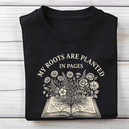 A folded black t-shirt with a cream-colored graphic print that reads 'MY ROOTS ARE PLANTED IN PAGES' above an illustration of an open book with flowers blooming from it, displayed on a white wood plank background.
