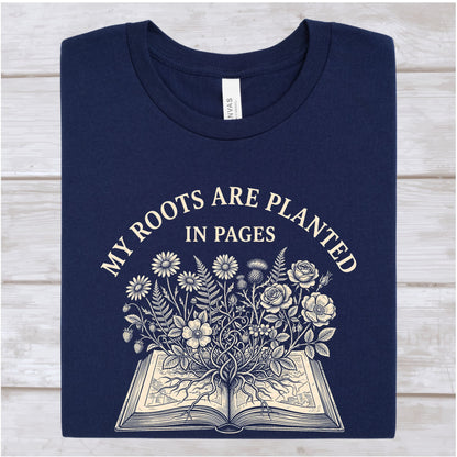 A navy blue crewneck t-shirt with a cream-colored graphic print that reads 'MY ROOTS ARE PLANTED IN PAGES' above an illustration of flowers growing from an open book, laid flat on a light wood background.