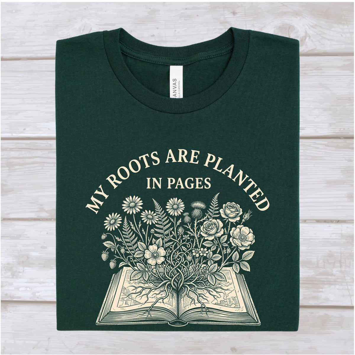 A dark forest green folded t-shirt with a cream-colored graphic print that reads 'MY ROOTS ARE PLANTED IN PAGES' above an illustration of an open book with flowers growing from the pages. The shirt is laid flat on a light-colored wooden plank background.