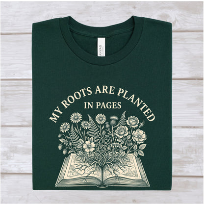 A dark forest green folded t-shirt with a cream-colored graphic print that reads 'MY ROOTS ARE PLANTED IN PAGES' above an illustration of an open book with flowers growing from the pages. The shirt is laid flat on a light-colored wooden plank background.