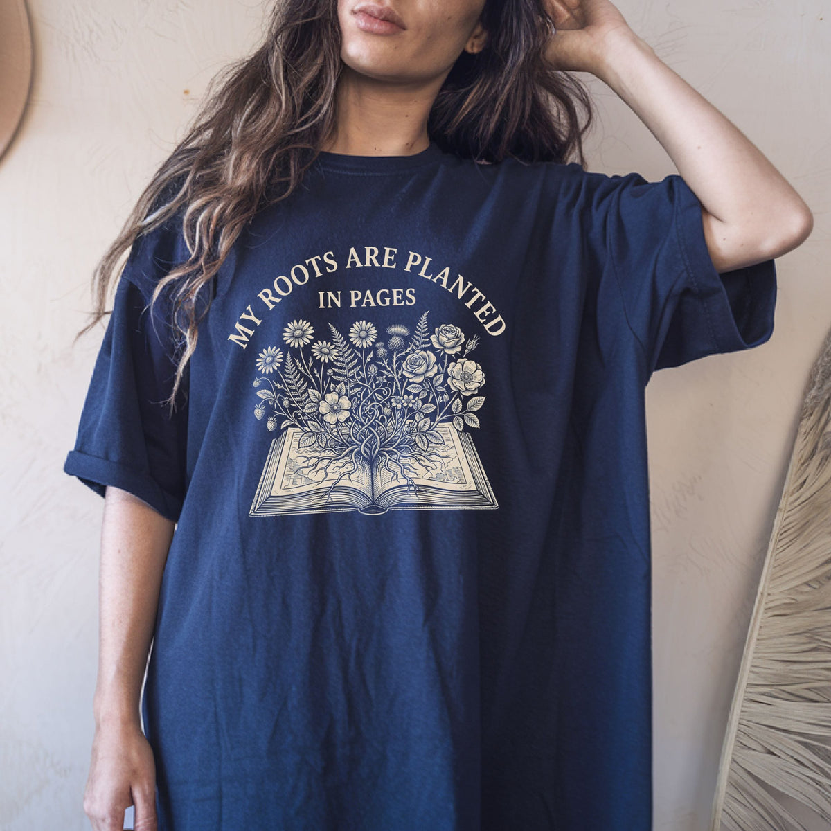 A woman wearing an oversized navy blue graphic t-shirt with a cream-colored print that reads 'MY ROOTS ARE PLANTED IN PAGES' above an illustration of an open book with flowers growing out of it.
