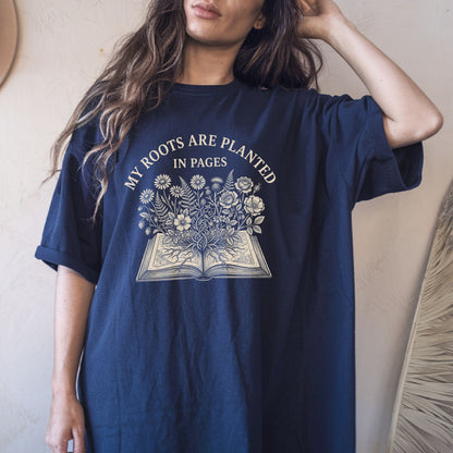 A woman wearing an oversized navy blue graphic t-shirt with a cream-colored print that reads 'MY ROOTS ARE PLANTED IN PAGES' above an illustration of an open book with flowers growing out of it.