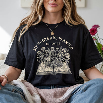 A woman wearing a black graphic t-shirt with a cream-colored print that reads 'MY ROOTS ARE PLANTED IN PAGES' above an illustration of an open book with flowers growing out of it. She is sitting indoors with a pink blanket on her lap.