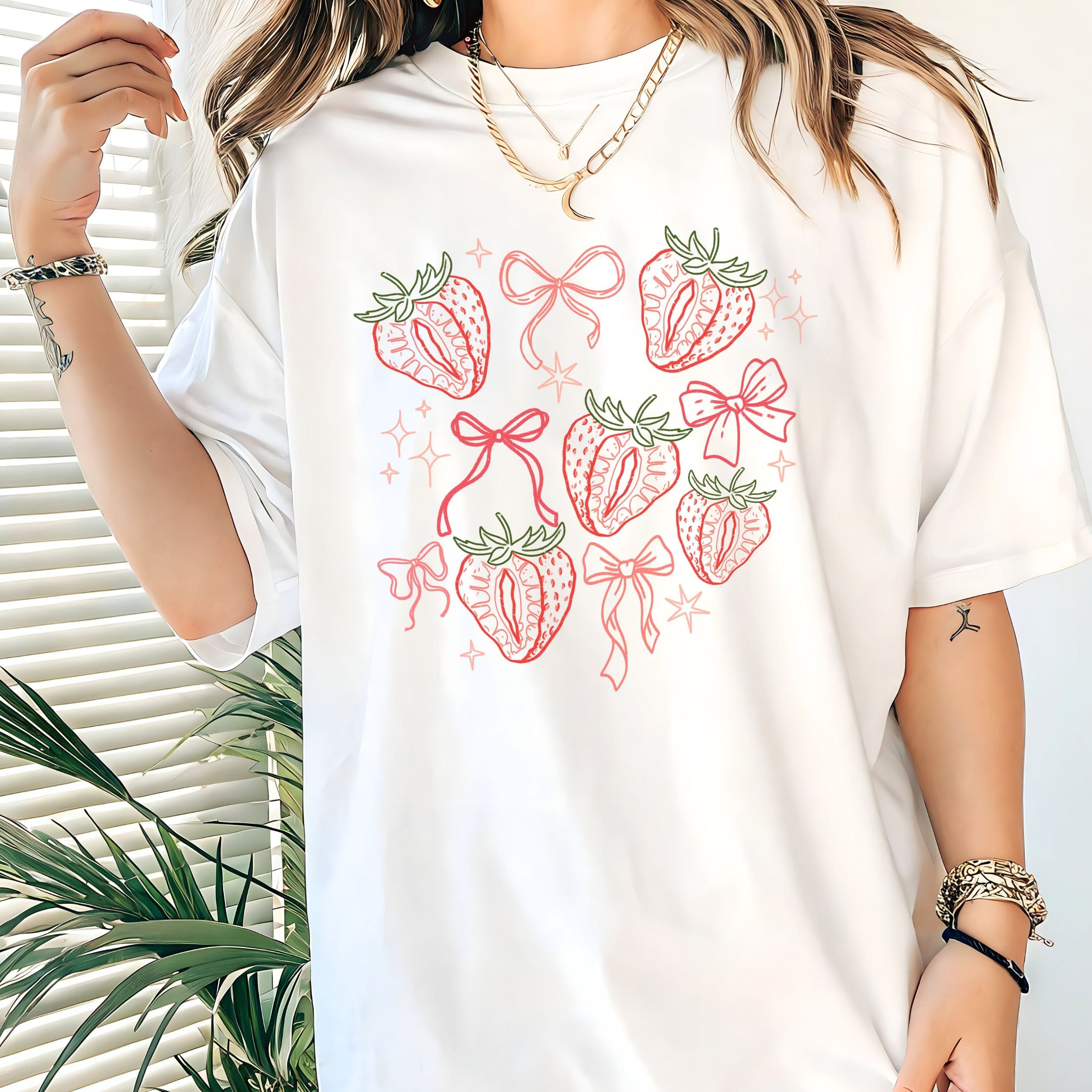 White t-shirt with strawberry and bow design worn by a person, with a plant in the background.