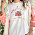 Person wearing a white t-shirt with 'Strawberry Festival' design, standing indoors.