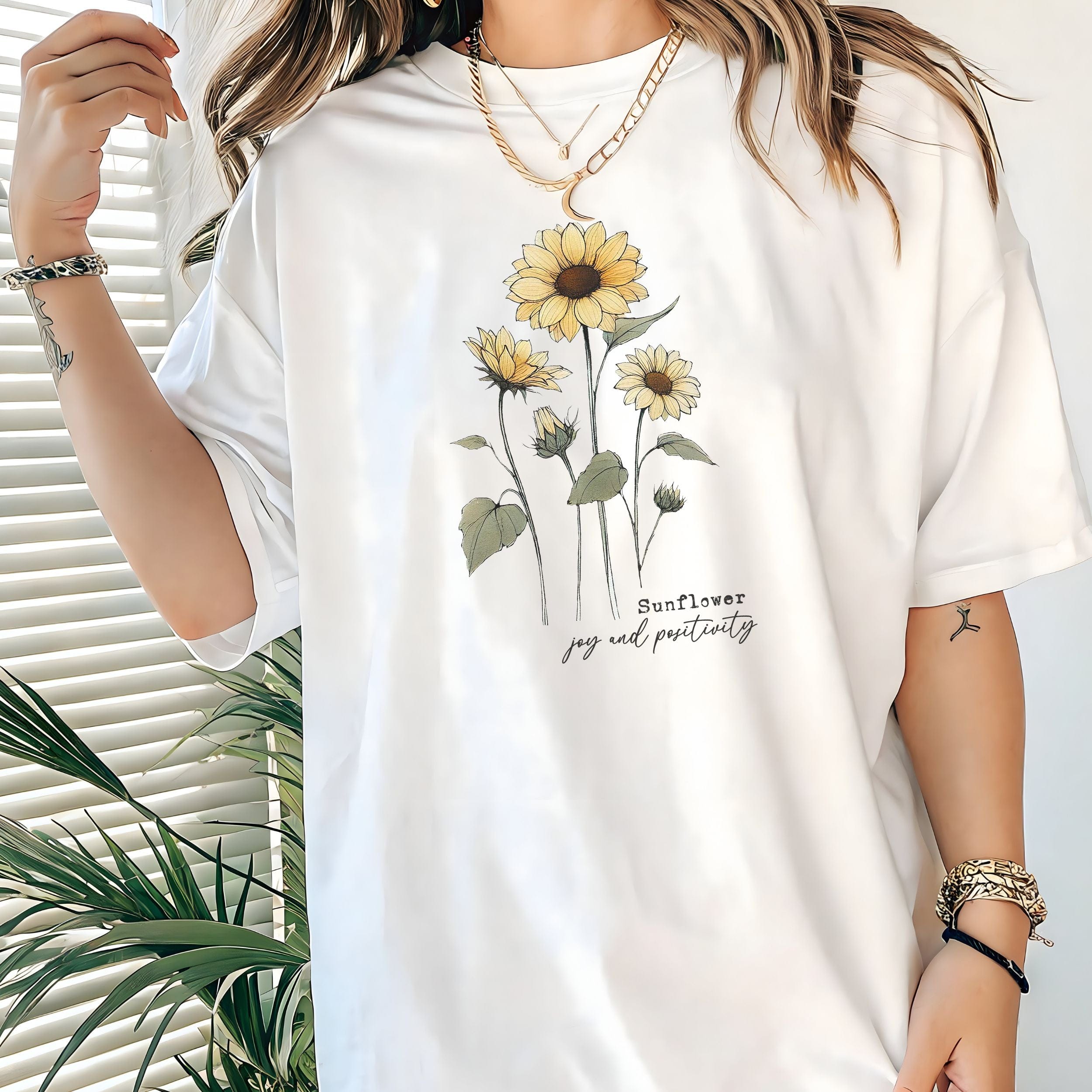 White t-shirt with sunflower design worn by a person, with a plant in the background.