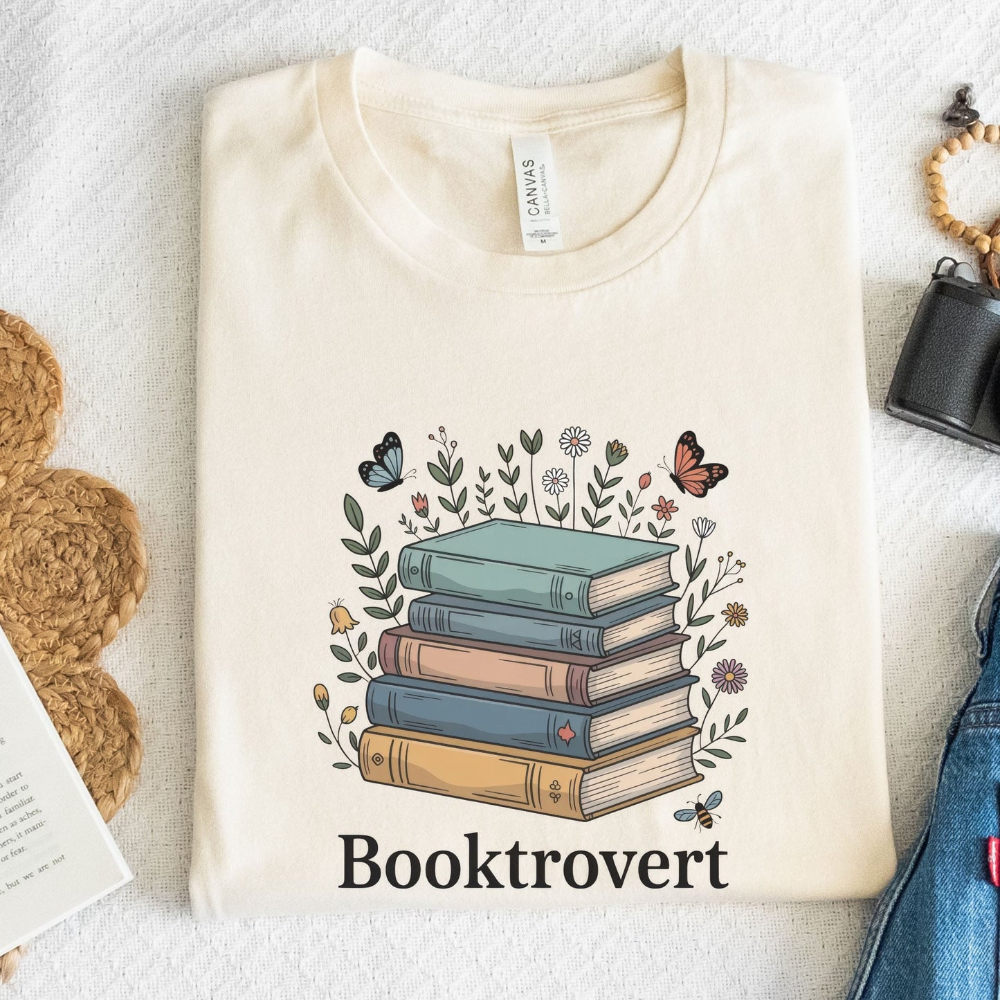 Beige t-shirt with a design of books and flowers, labeled 'Booktrovert', on a textured surface.