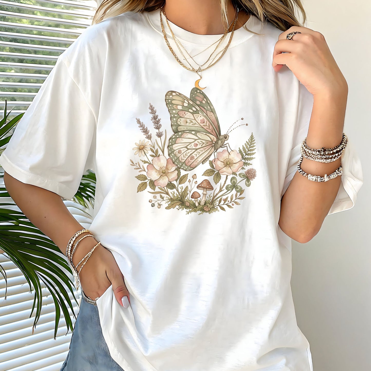 White t-shirt with a butterfly and floral design worn by a person, with a plant in the background.