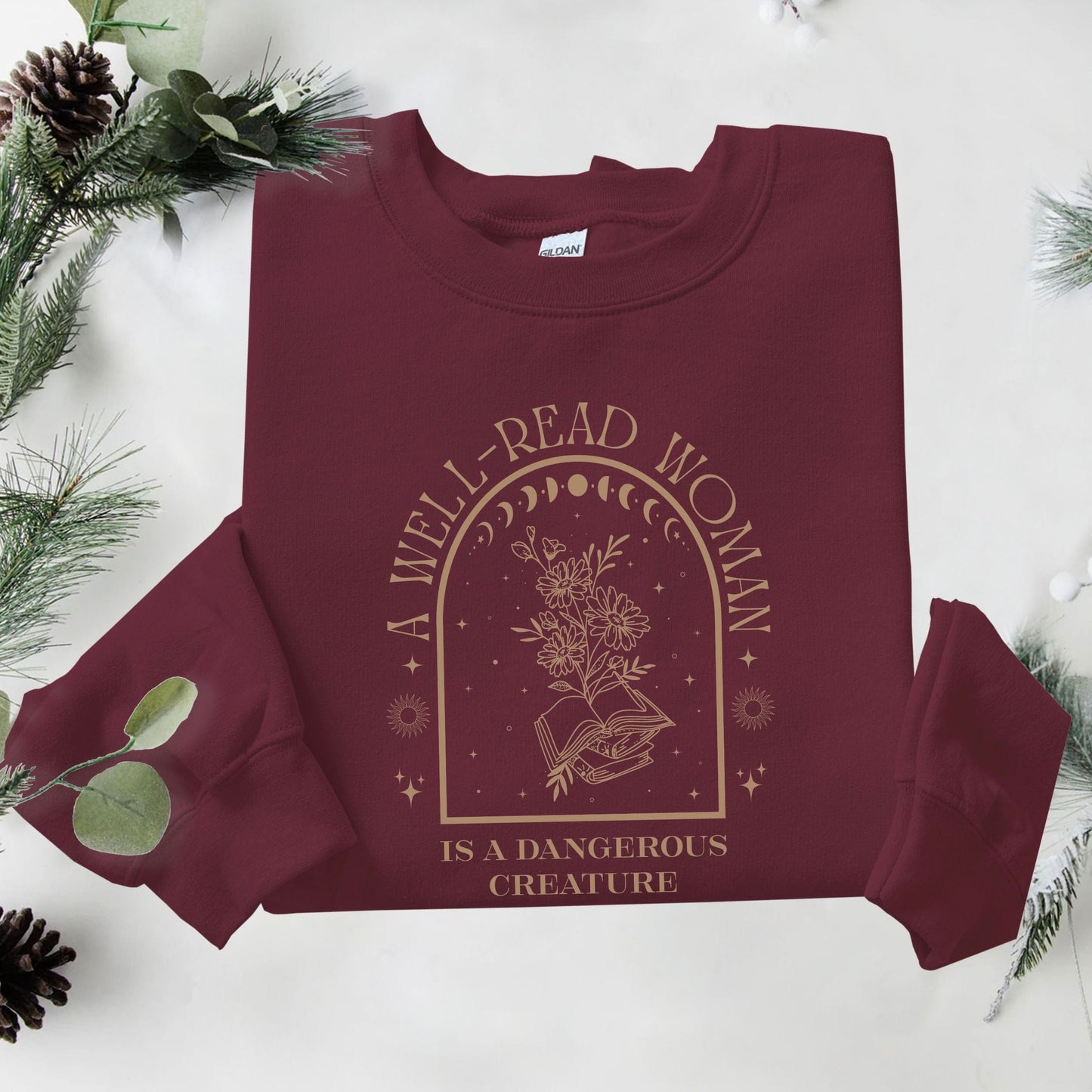 Maroon sweatshirt with 'A Well-Read Woman is a Dangerous Creature' text and design on a white background with greenery.