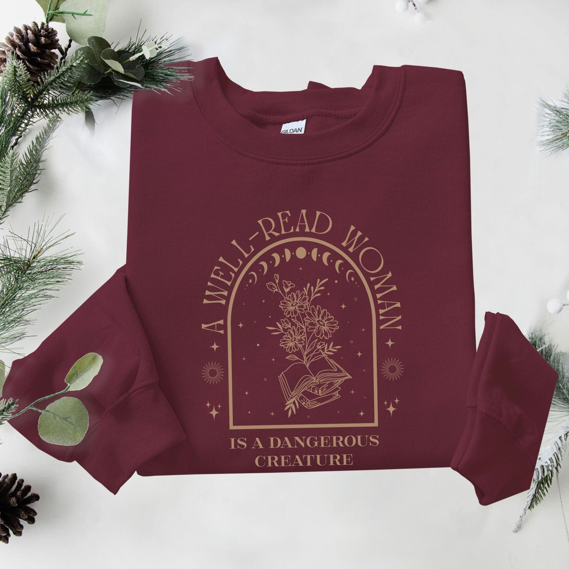 Maroon sweatshirt with 'A Well-Read Woman is a Dangerous Creature' text and design on a white background with greenery.