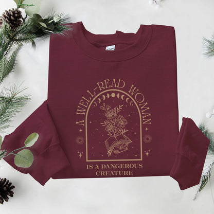 Maroon sweatshirt with 'A Well-Read Woman is a Dangerous Creature' text and design on a white background with greenery.
